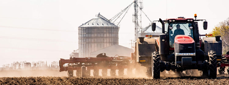 Tractor Driving through Field with Silos in Background